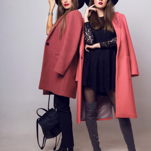 Full height   studio image of two fashionable sexy woman in casual trendy spring coat,  boots with heels , black hat, stylish hand bag.